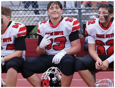 Two people wearing white and red football jerseys with black pants are sitting on a bench, one holding a helmet and the other with black face paint. Description generated by AI