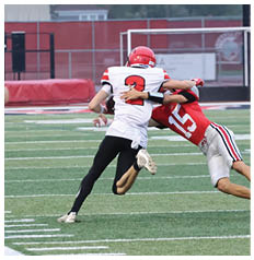 Two people in football uniforms are engaged in a tackle on a football field while a person in black athletic clothing watches from the sideline. Description generated by AI