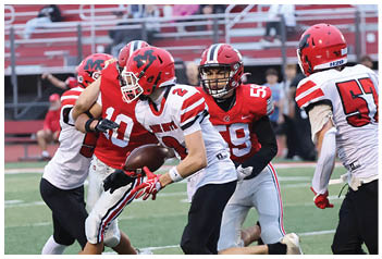 People in football uniforms are actively engaged in a game on a field with bleachers and spectators in the background. Description generated by AI