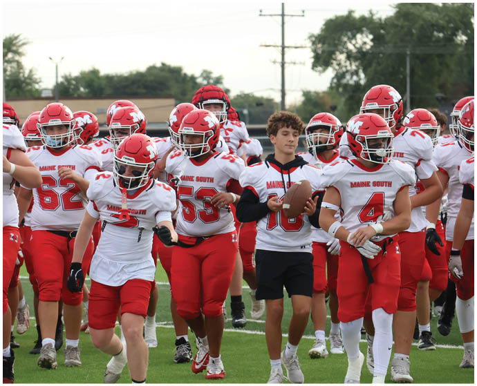 A group of people wearing red and white football uniforms with helmets are walking together on a grassy field, with one person holding a football and wearing a jersey over a black hoodie and shorts. Description generated by AI
