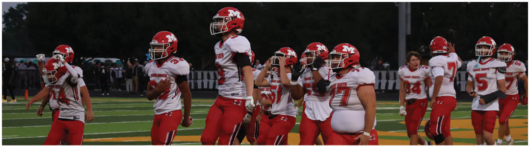 People wearing red and white football uniforms are walking on a green field with yard lines visible. Description generated by AI