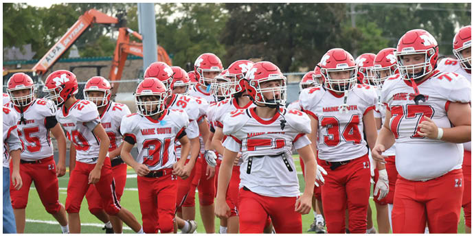 A group of people wearing red and white football uniforms and helmets are gathered on a grassy field with trees and construction equipment in the background. Description generated by AI