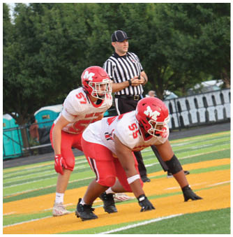 Two teams of football players in red and black uniforms are lined up on a field with a referee standing behind them. Description generated by AI