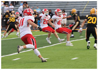 People in red and white uniforms are running on a football field toward a person in a black and yellow uniform during a game with empty bleachers in the background. Description generated by AI