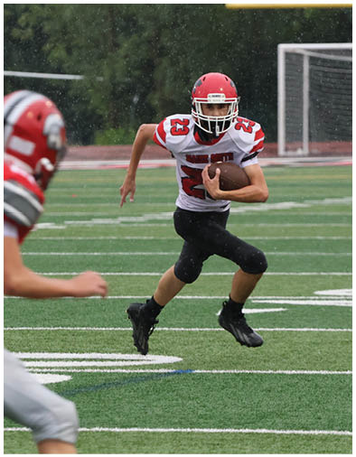 A person wearing a red helmet and white football jersey with the number 23 is running on a football field while holding a football, with another player partially visible in the foreground. Description generated by AI