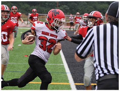 A person wearing a red helmet and white jersey with the number 32 is running with a football while another person in a red helmet and gray pants reaches out to block them on a football field. Description generated by AI