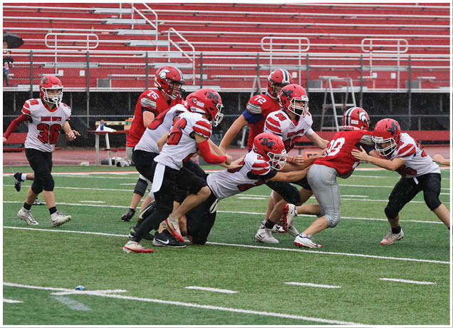 Several people wearing football helmets and uniforms are actively engaged in a tackle on a green field with red bleachers in the background. Description generated by AI