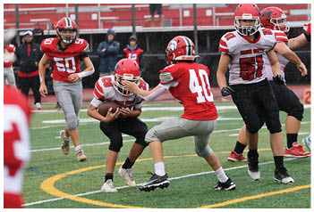 People wearing red and white football uniforms are playing on a green field with yellow markings, with one person holding the ball while others are running and preparing to tackle. Description generated by AI