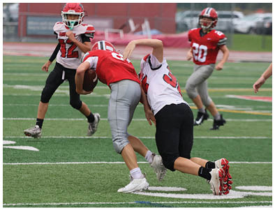 People wearing sports uniforms are playing football on a grassy field with one person holding the ball while others attempt to tackle. Description generated by AI