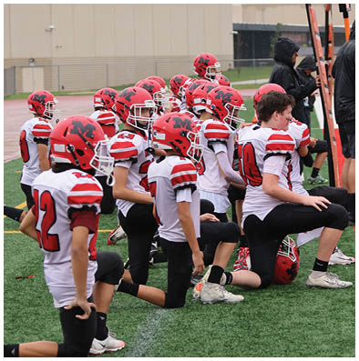 A group of people wearing red helmets and white sports jerseys with red and black accents are sitting closely together on a field, likely during a football game or practice. Description generated by AI