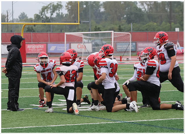 A group of people wearing red helmets and white sports jerseys with black pants are kneeling on a football field near a goalpost. Description generated by AI