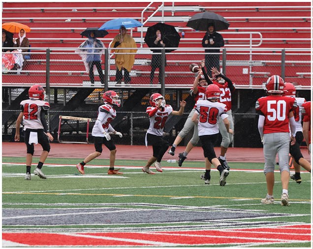 Children playing football on a field with some people in the background holding umbrellas and sitting or standing in front of empty red bleachers. Description generated by AI