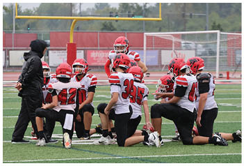 Three people wearing red helmets and white football uniforms are on a sports field, with one person pointing in a direction. Description generated by AI