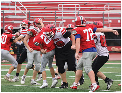 Several people wearing football helmets and uniforms are engaged in a play on a football field with red bleachers in the background. Description generated by AI