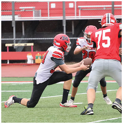 Three people wearing red helmets and sports uniforms are playing football on a field, with one person holding the ball and another preparing to receive it. Description generated by AI