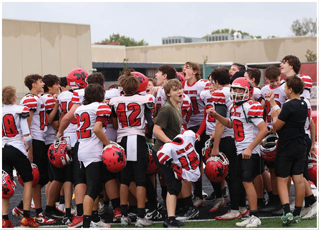 A group of people wearing red and white football uniforms are gathered closely together outdoors, some cheering and one person in a green shirt appears to be shouting. Description generated by AI