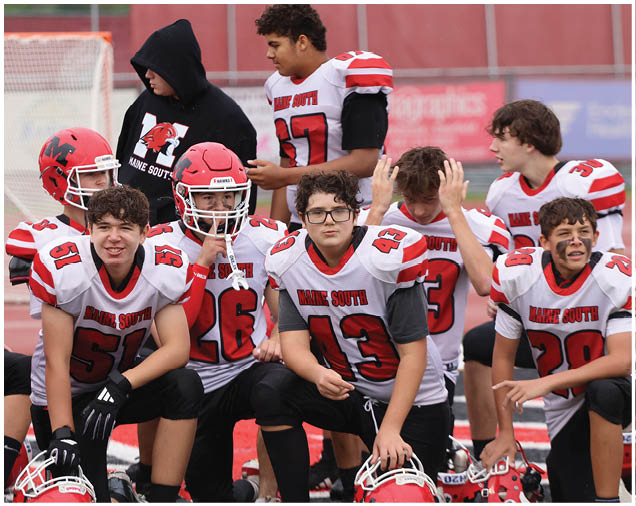 A group of people wearing white and red football uniforms are gathered together on a field, some sitting and some standing, with a few holding helmets. Description generated by AI