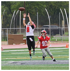 Two people in football uniforms are on a field with one jumping to catch a football while the other runs nearby. Description generated by AI