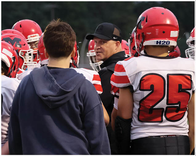 A group of people wearing red and white football uniforms and helmets are gathered around a person in a black cap and jacket who appears to be giving instructions. Description generated by AI