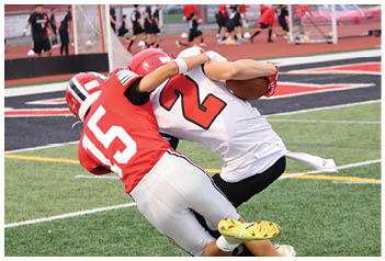 Two people in football uniforms are engaged in a tackle on a grassy field during a game. Description generated by AI