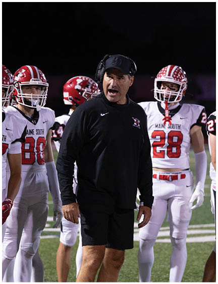 A coach wearing a headset and black sportswear is speaking energetically to a group of football players in white and red uniforms on a field at night. Description generated by AI