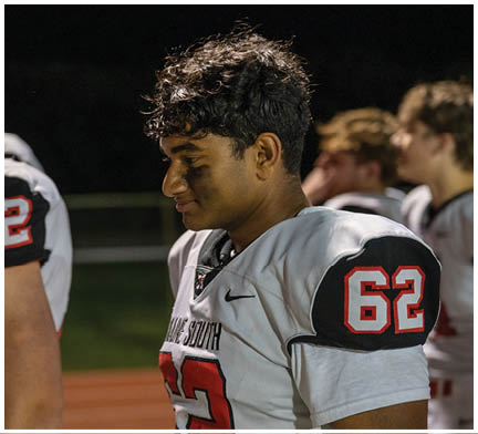 A person wearing a football uniform with the number 62 and black face paint stands on a field at night next to another person in a uniform with the number 52. Description generated by AI