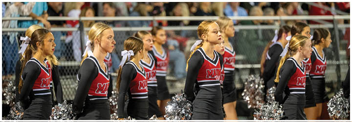 A group of people in matching cheerleading uniforms stand in a row outdoors with a crowd and fence in the background. Description generated by AI