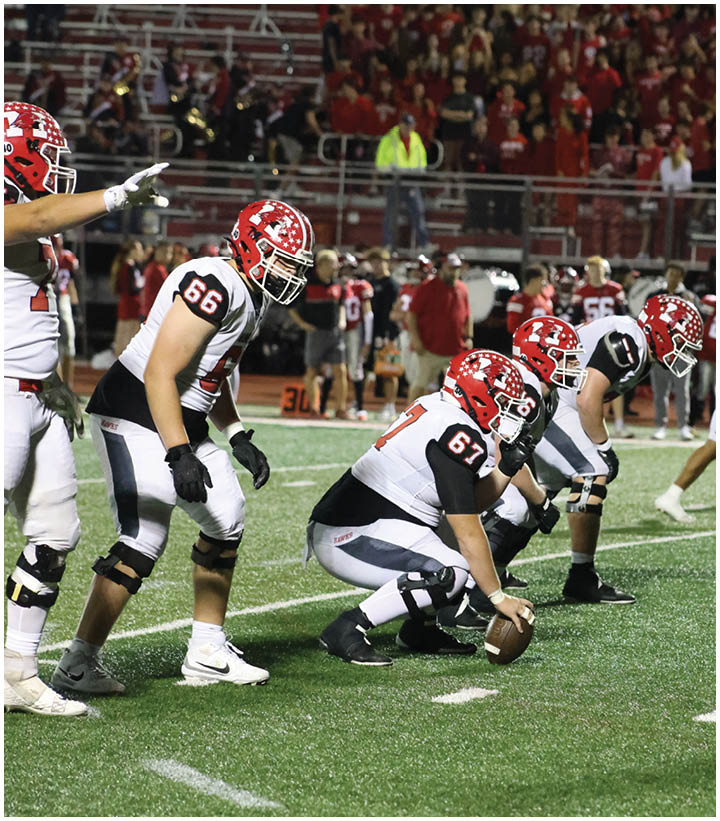 A group of football players in red helmets and white and black uniforms are lined up on a green field preparing for a play during a game. Description generated by AI
