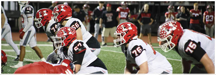 Several football players in red and white uniforms are lined up on a field preparing for a play during a game. Description generated by AI