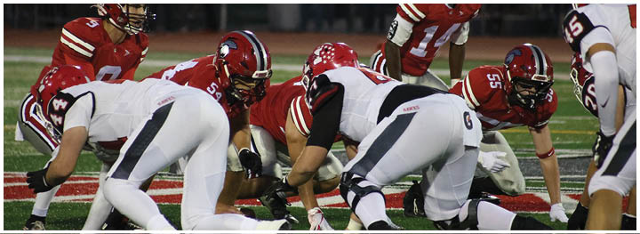 People in red and white football uniforms are lined up on a field preparing for a play during a game. Description generated by AI