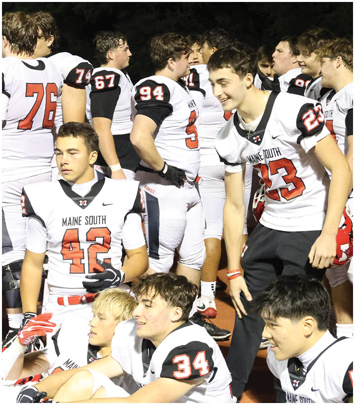 A group of people wearing white football uniforms with red and black accents are gathered on a field, some standing and some sitting, with one person holding a red helmet. Description generated by AI