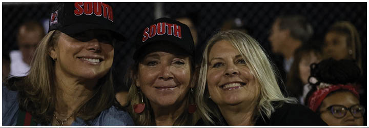 Three adults smiling closely together at an outdoor event during nighttime, with two wearing black caps that say “SOUTH." Description generated by AI