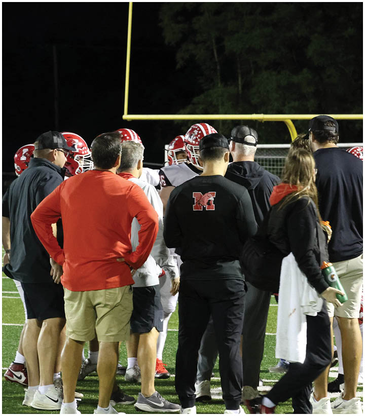 Several people in casual and sports clothing stand on a football field near players wearing helmets and uniforms under stadium lights at night. Description generated by AI