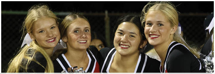 Four people wearing cheerleading uniforms with pom-poms smile closely together in an outdoor setting. Description generated by AI