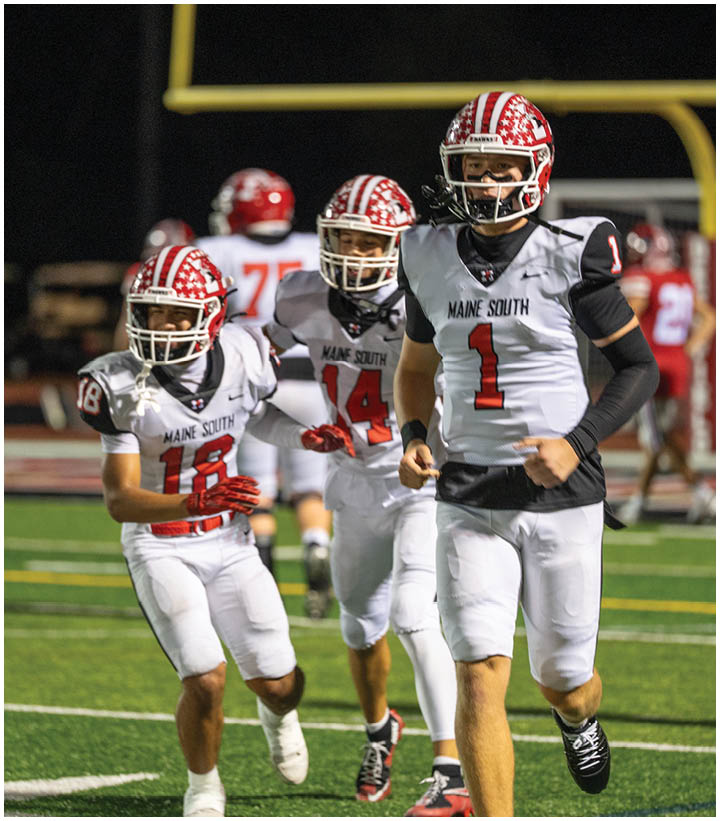 Three people in white football uniforms with red helmets and gloves are running on a green field during a night game. Description generated by AI