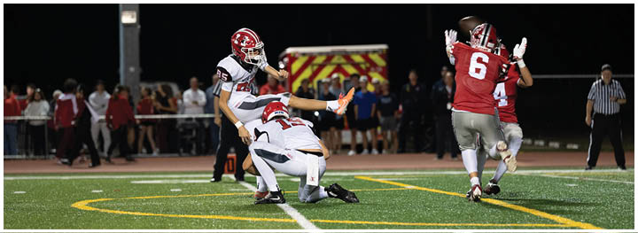 A person in a red sports uniform is running on a football field while two people in white uniforms are attempting to tackle them during a nighttime game. Description generated by AI