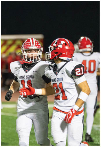 Two people wearing white football uniforms with red helmets and gloves are on a field, appearing to communicate during a game. Description generated by AI