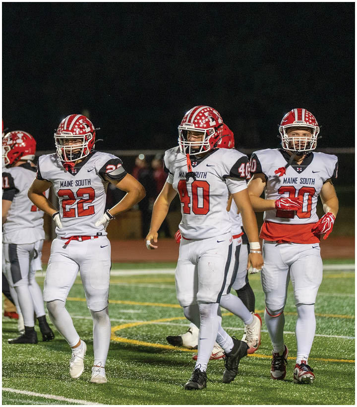 Four people in football uniforms stand on a field at night with three wearing white jerseys labeled “Maine South" and one in a red jersey. Description generated by AI