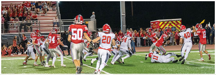 People in red and white football uniforms are actively playing on a well-lit field with spectators in the stands and an emergency vehicle in the background. Description generated by AI