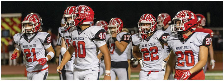 A group of people wearing red helmets and white sports uniforms with “Maine South" on the front are walking together on a sports field at night. Description generated by AI