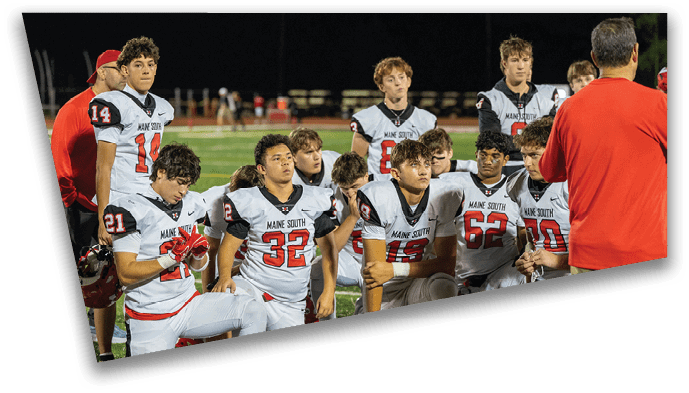 A group of people wearing football uniforms with “Maine South" on the jerseys are kneeling on a field while listening to a person in a red jacket. Description generated by AI
