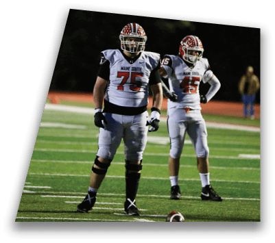 Two people in football uniforms with helmets stand on a field at night, one wearing number 75 and the other number 12. Description generated by AI