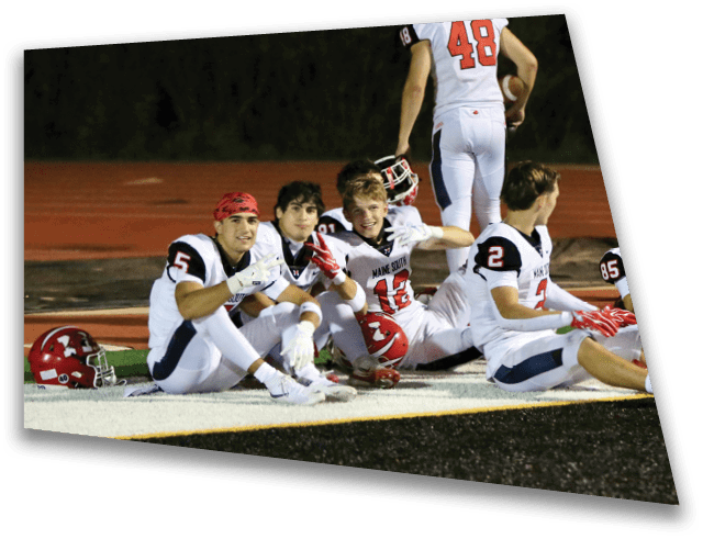 Four people in football uniforms are sitting and lying on the field near the sideline, with one person standing and holding a football in the background. Description generated by AI