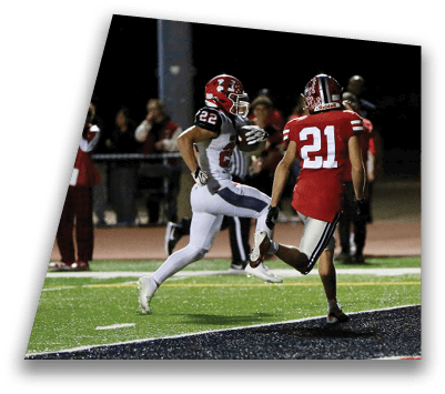 Two people in football uniforms are running on a field near the end zone, with one holding a football. Description generated by AI