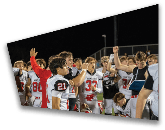 A group of people in football uniforms are gathered together at night, with one person raising a fist and another wearing an arm sling. Description generated by AI