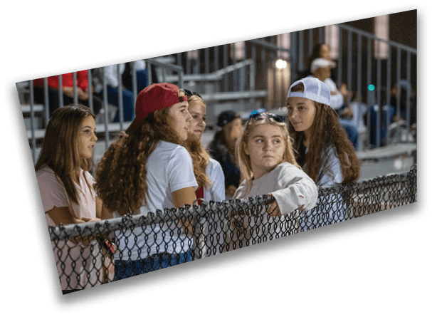 A group of five young people wearing casual clothing and hats are sitting and standing near a chain-link fence in a stadium or bleacher setting at night. Description generated by AI