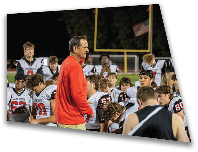 A group of people wearing football uniforms are gathered on a field while a person in a red jacket appears to be speaking to them. Description generated by AI