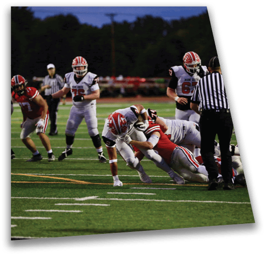 People in football uniforms are engaged in a tackle on a field with a referee nearby and other players watching. Description generated by AI