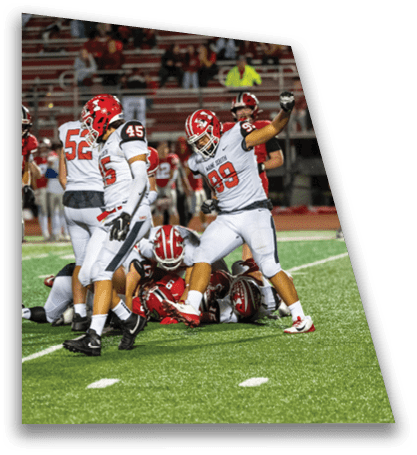 Several football players in white and red uniforms are gathered on a green field with one player raising a fist in celebration while others are on the ground. Description generated by AI