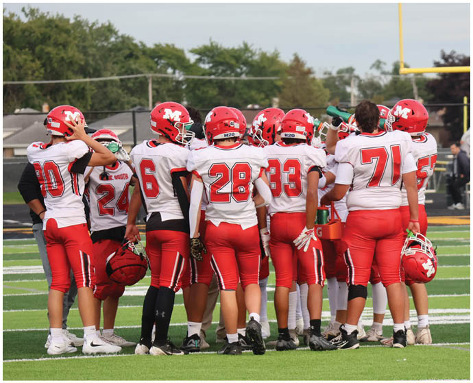 A group of people wearing red and white football uniforms are gathered on a field, some holding helmets and water bottles. Description generated by AI